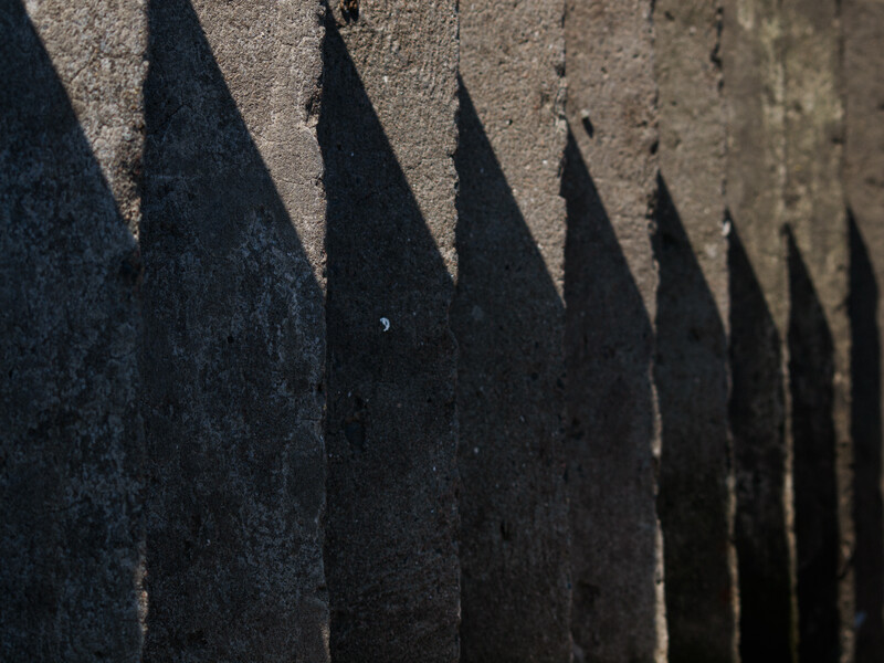 Sharp metal fence spikes casting long shadows on a textured concrete surface in bright sunlight.