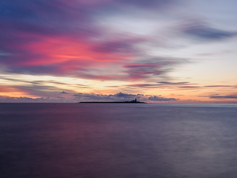 Seascape at sunset with pink and purple clouds reflected in calm water and a distant silhouetted island.