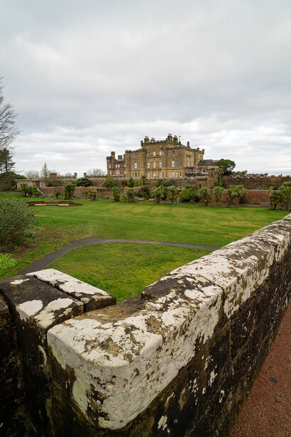 Stone wall in foreground with manicured lawn and grand castle structure beyond under overcast sky.