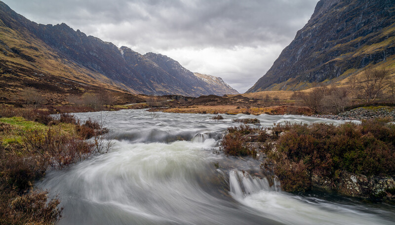 River with silky water flowing through a mountain valley under stormy skies.