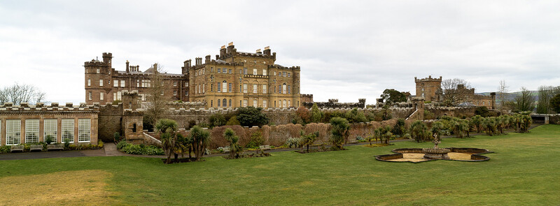 Stone castle with manicured grounds, formal gardens, and rolling landscape under cloudy sky.