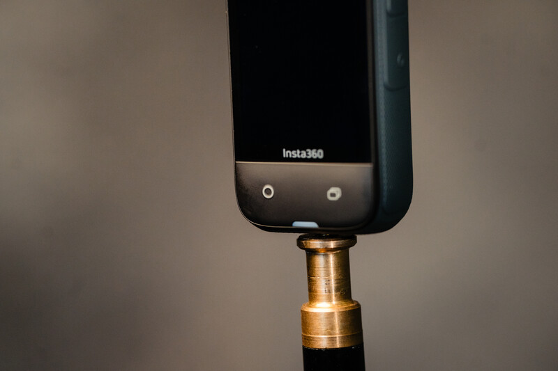Insta360 action camera mounted on a brass extension pole against a neutral beige background.