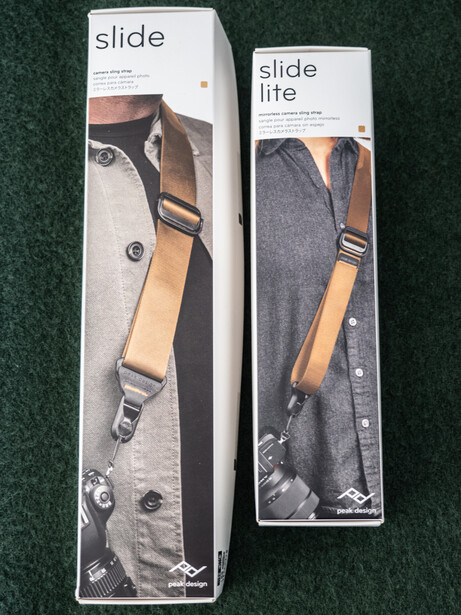 Two packaged camera shoulder straps with wooden handles displayed side by side on green fabric.