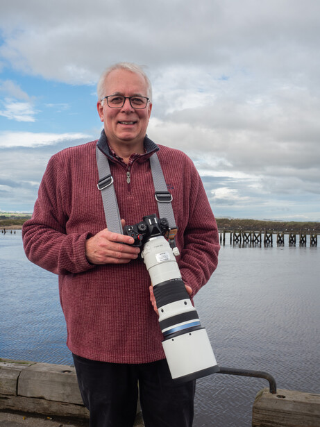 Photographer holding a large telephoto lens while standing at a waterfront location.