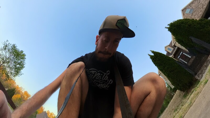 Man in black t-shirt and tan cap looking down at camera with wide-angle perspective on residential street.