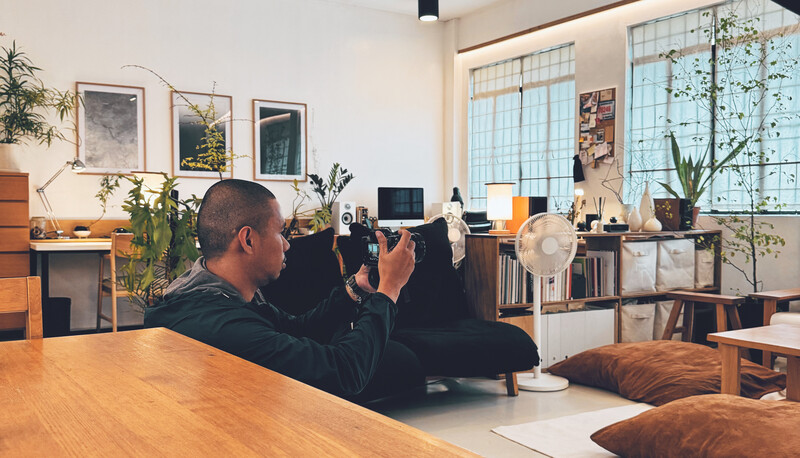 Photographer holding camera up to shoot in a bright, plant-filled studio space with large windows.