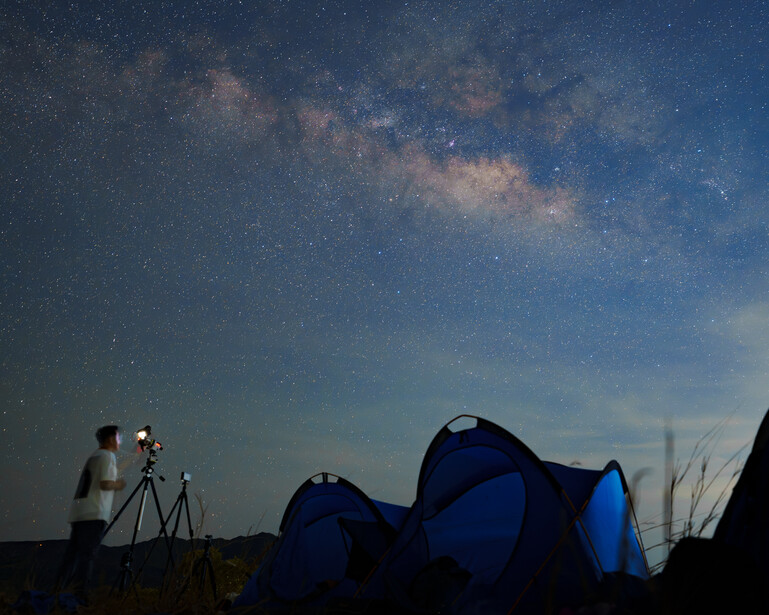 Silhouetted photographer at tripod capturing Milky Way galaxy above illuminated blue camping tents.