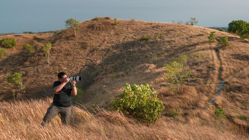 Photographer in dark clothing shooting with a telephoto lens on a hillside with sparse vegetation and dry grass.
