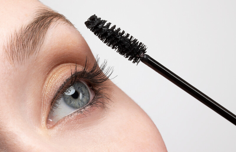 Close-up of a mascara wand being applied to an eye with warm-toned eyeshadow and defined lashes.