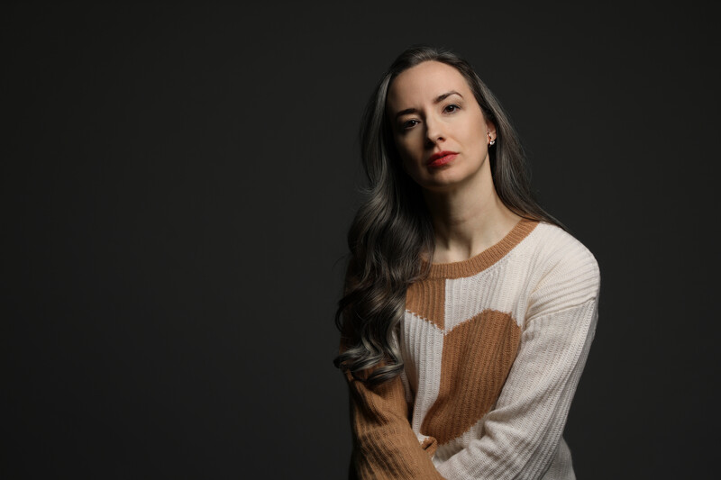 Woman with long dark hair wearing a cream and brown color-block ribbed sweater against a dark background.