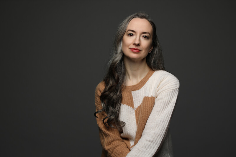 Woman with long dark hair wearing a cream and caramel colorblock sweater against a dark gray background.