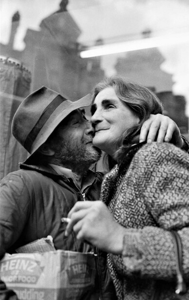 Man in hat and woman in close embrace, shot in black and white with blurred urban architecture behind them.