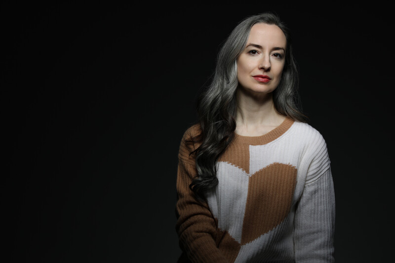 Woman with long dark hair wearing a brown and white color-block sweater against a black background.