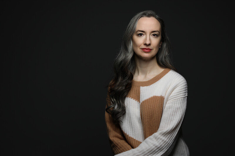 Woman with long dark hair wearing a cream and tan color-blocked sweater against a dark background.