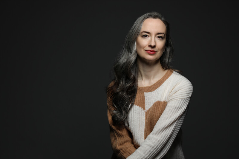 Woman with long dark hair wearing a brown and cream striped sweater against a black background.
