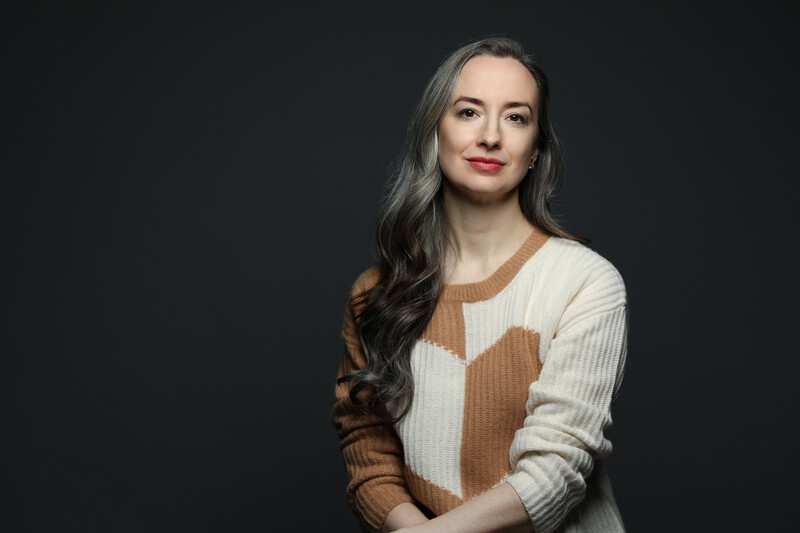 Woman with long dark hair wearing a cream and brown geometric patterned sweater against a dark background.