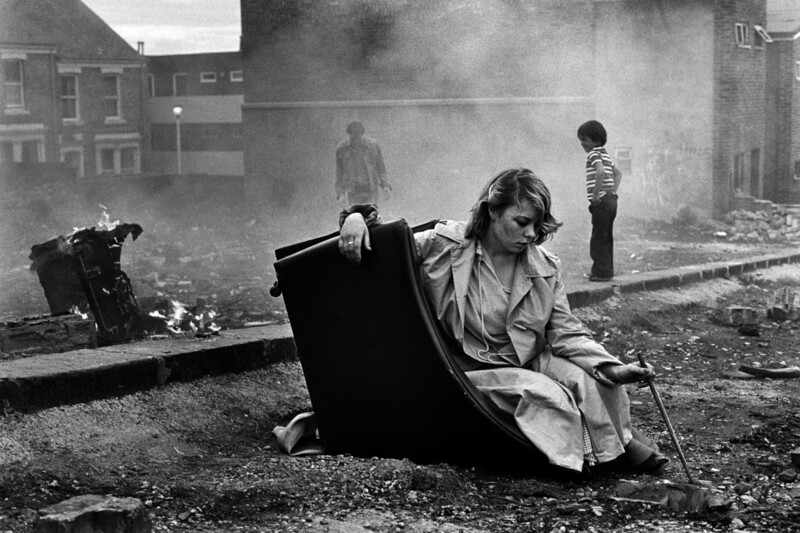 Woman crouching amid urban rubble and smoke, with figures visible in hazy background.