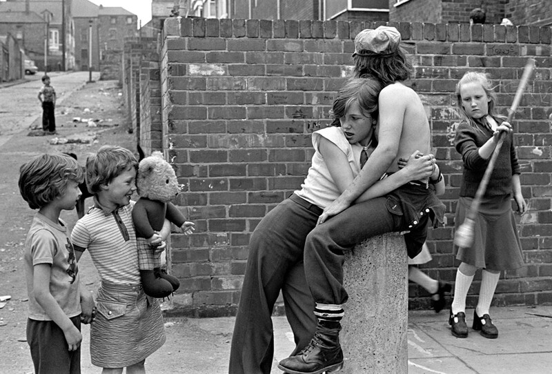Woman holding a young child while surrounded by other children against a brick wall.