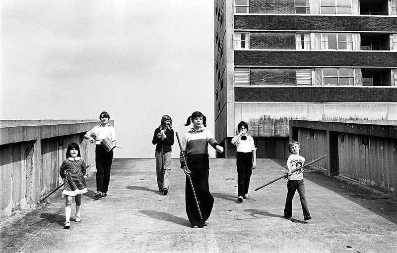 Children and adults playing with a large tire on a concrete rooftop beside a modernist apartment building.