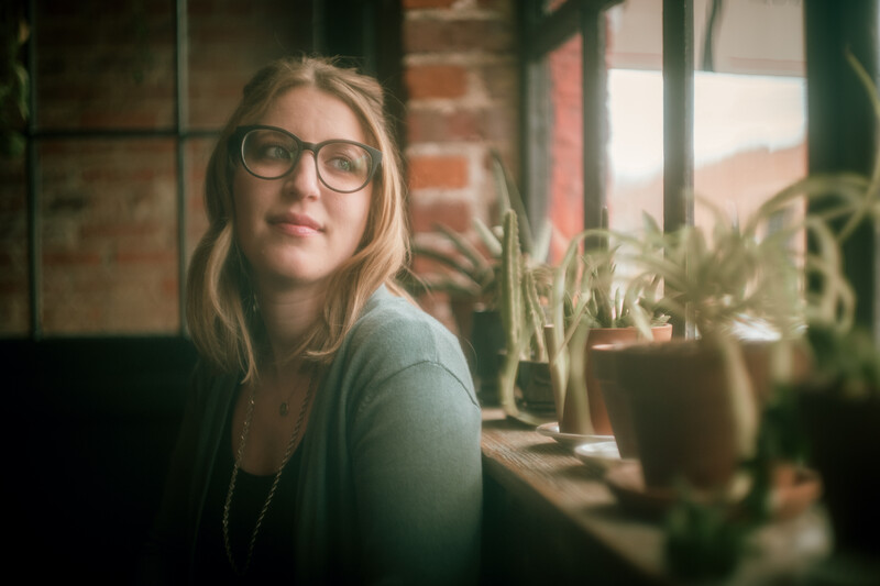 Woman wearing glasses gazing to the side near windowsill plants in warm, natural light.