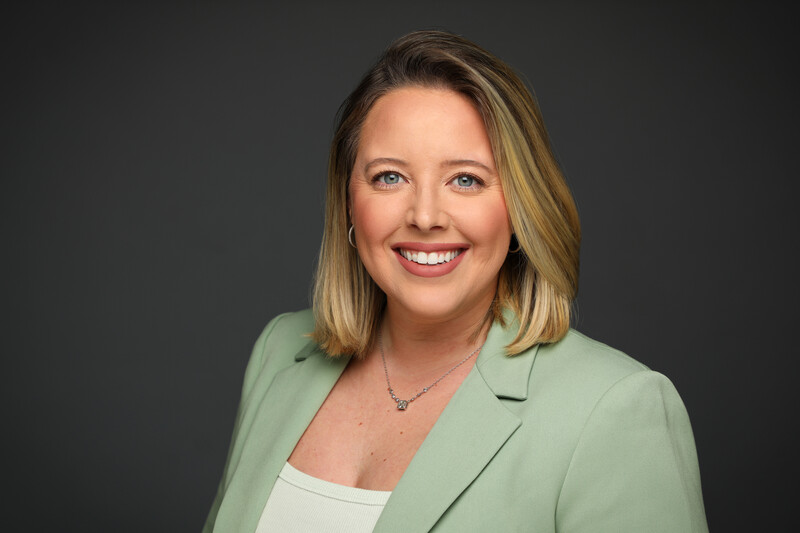 Professional headshot of a woman with shoulder-length blonde hair wearing a sage green blazer over a white top.