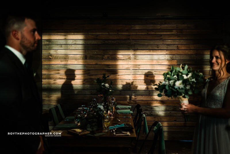 Couple at an indoor reception with warm wooden wall backdrop and ambient lighting casting shadows.