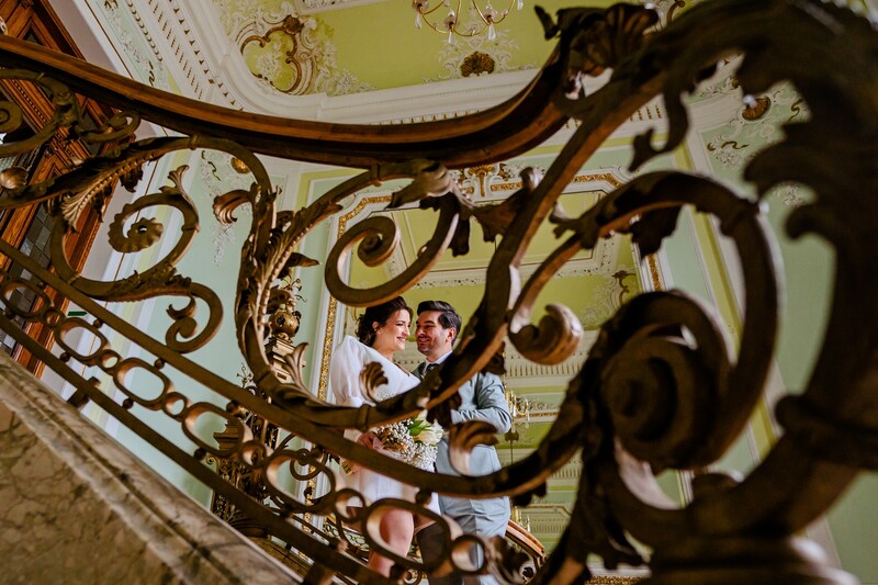 Couple on ornate spiral staircase viewed through decorative iron railing with baroque scrollwork.