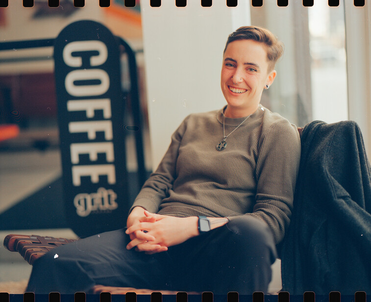 Smiling person sitting against a wall with a coffee shop sign visible in the blurred background.