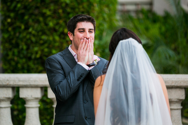 Groom in dark suit covers his mouth in surprise while bride in white veil stands before him.