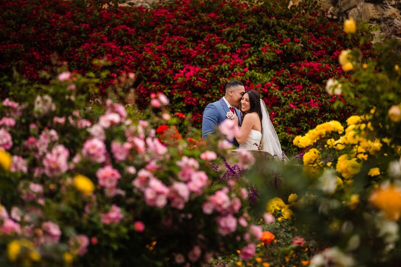 Couple kissing in a flower garden with vibrant pink, red, and yellow blooms surrounding them.