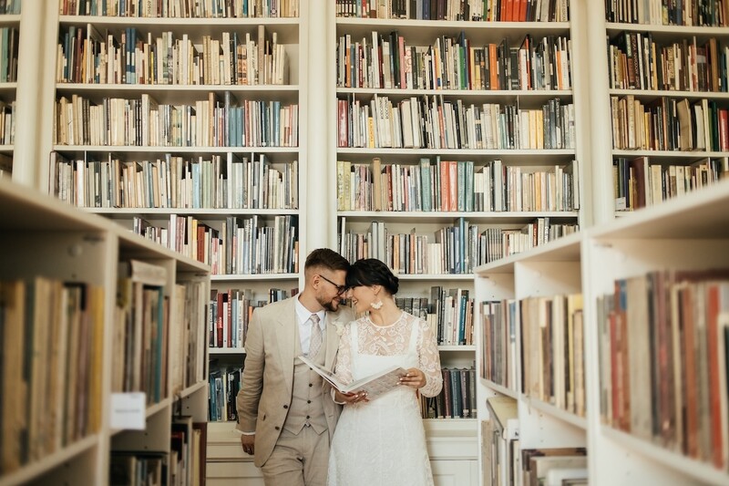 Couple in formal attire reading together in a library aisle lined with bookshelves.