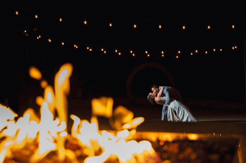 Couple kissing by a firepit at night with string lights overhead.