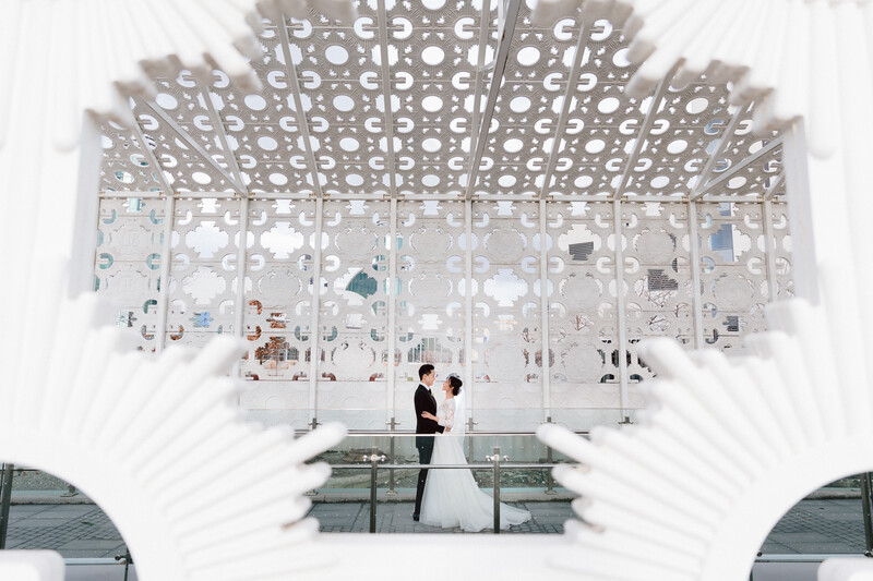 Bride and groom standing on a glass platform beneath an ornate geometric ceiling structure in a minimalist white venue.