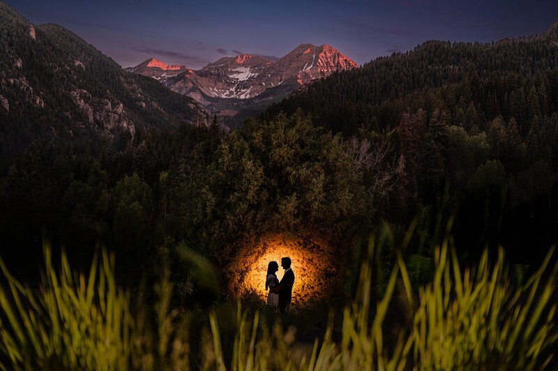 Silhouetted couple illuminated by warm light in a grassy meadow, with snow-capped mountains glowing at twilight.