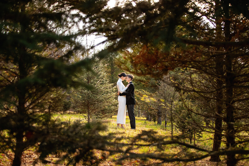 Bride and groom standing together in a forested clearing, backlit by golden sunlight filtering through trees.