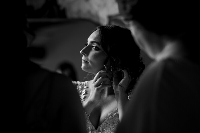 Bride in profile wearing lace wedding dress, hands near her neck in contemplative pose. Black and white portrait photography.