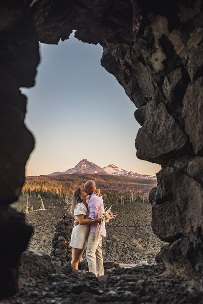 Couple standing in a natural rock archway overlooking a volcanic mountain landscape at golden hour.