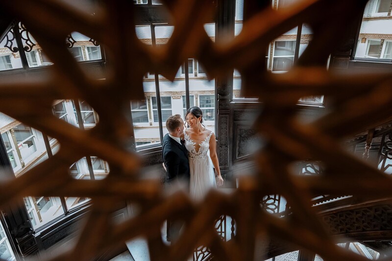 Bride and groom standing together by large windows, photographed through an ornate wooden screen in soft natural light.