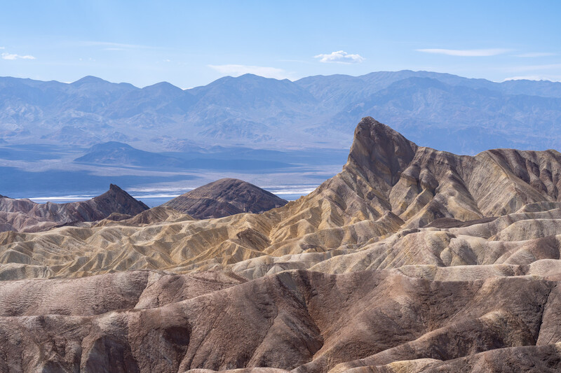 Eroded badlands terrain with sharp peaks and ridges in warm earth tones, with a vast valley and mountains visible under blue sky.