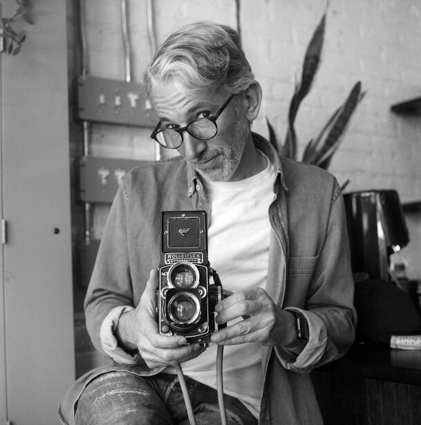 Photographer holding a twin-lens reflex camera, wearing glasses and cardigan over white shirt.
