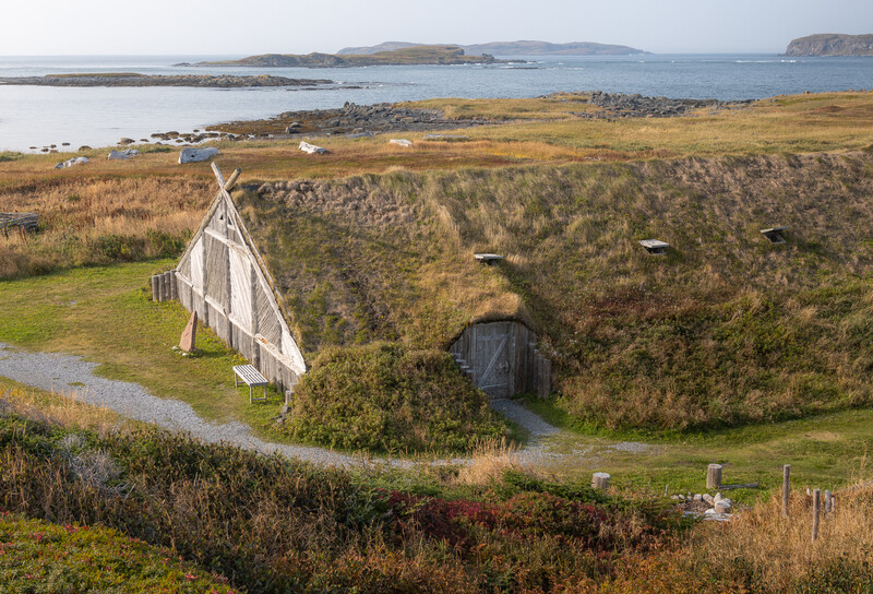 Turf-roofed dwelling beside a grassy hillside with coastal landscape and islands visible in the distance.