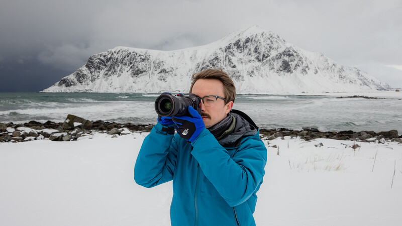 Photographer in blue jacket holding telephoto lens toward snow-capped mountain across frozen coastal waters.