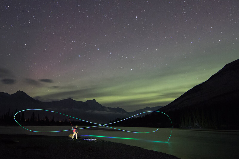 Light painter creates glowing infinity symbol on dark beach at night with starry sky and mountains.
