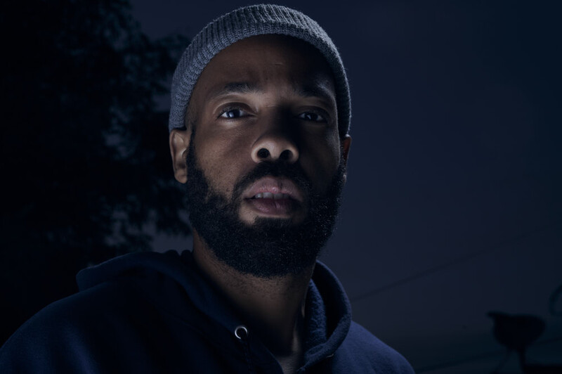 Man wearing gray beanie and dark hoodie against dark background, looking upward with serious expression.