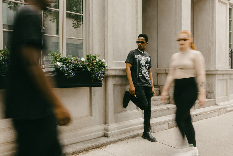 Man in sunglasses and graphic t-shirt walking past classical architecture with blurred pedestrians.