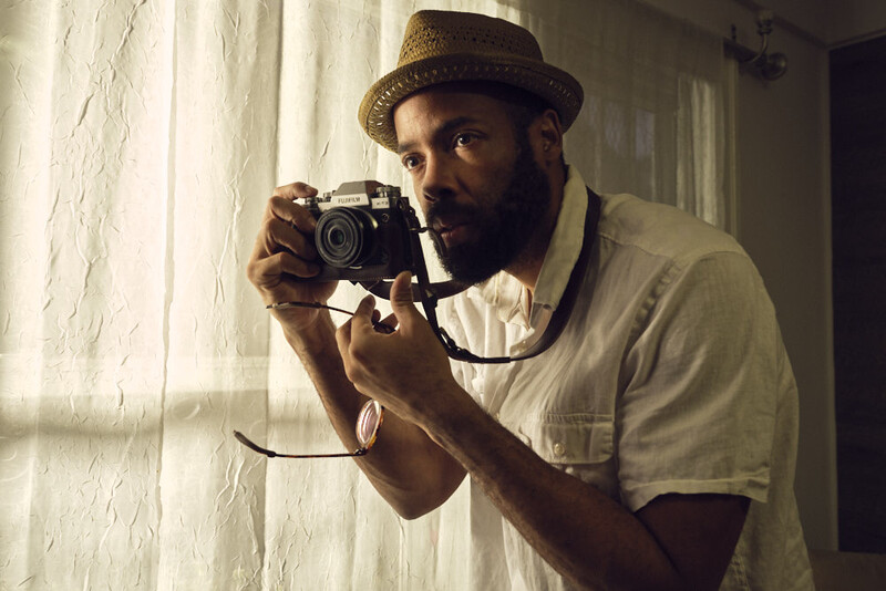 Photographer holding vintage rangefinder camera up to his eye, composing a shot indoors near a window.