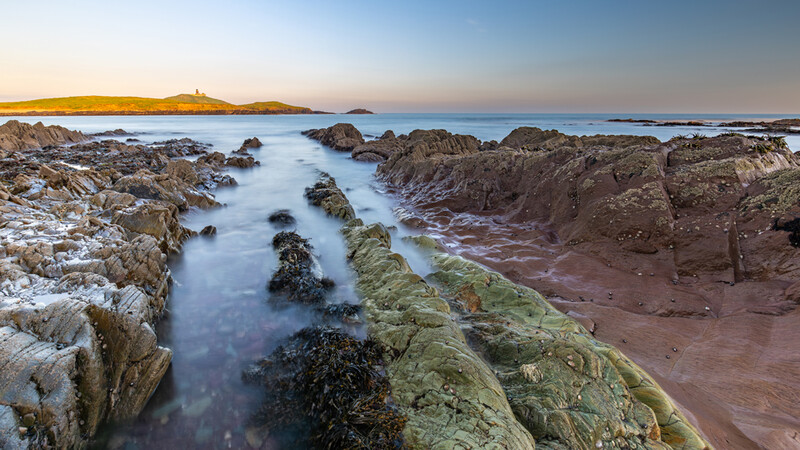 Rocky coastline at twilight with moss-covered stone formations and smooth water created by long exposure.