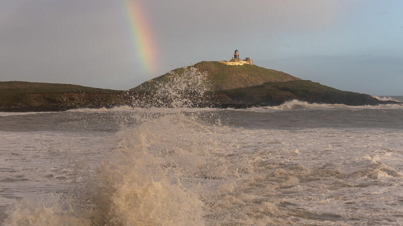 Ocean waves crash on a beach with a grass-covered headland and lighthouse silhouetted against stormy skies and a rainbow.