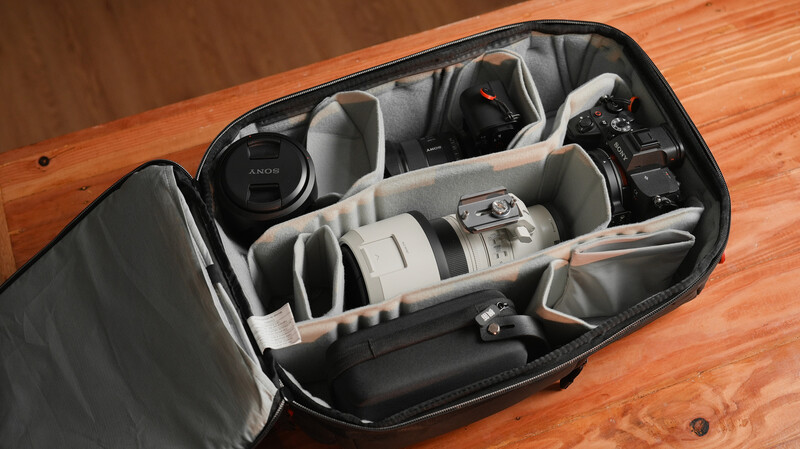 Camera bag interior with customizable gray dividers holding a camera body, white telephoto lens, and accessories.