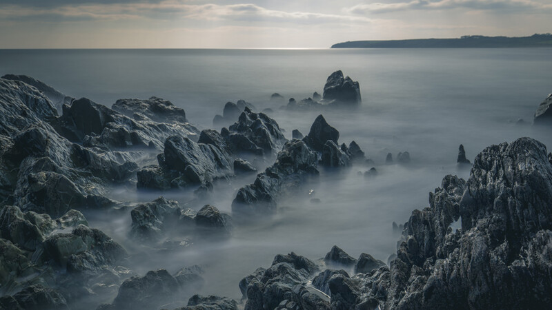Rocky coastline shrouded in heavy mist with jagged stone formations piercing through fog over calm water.